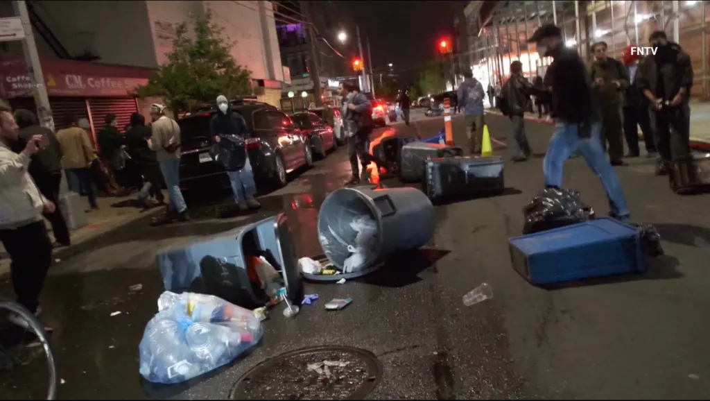Protesters gather on a street at night, with trash cans overturned and debris scattered on the ground.