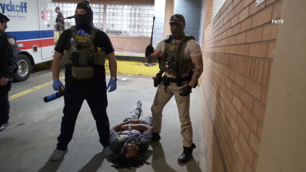 Two ICE officers, dressed in tactical gear and face coverings, stand over a man lying on the ground, with an ambulance visible in the background.