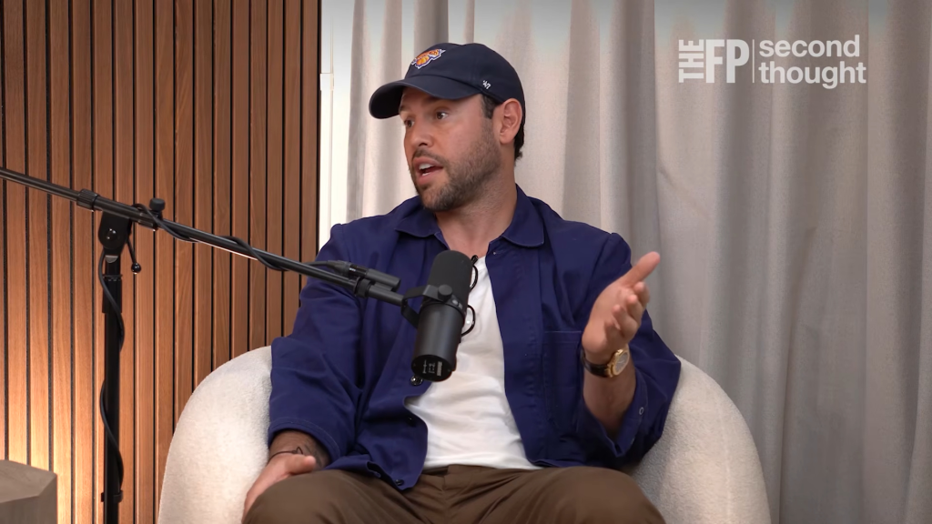 Scooter Braun in a navy cap and jacket, seated and speaking into a microphone.