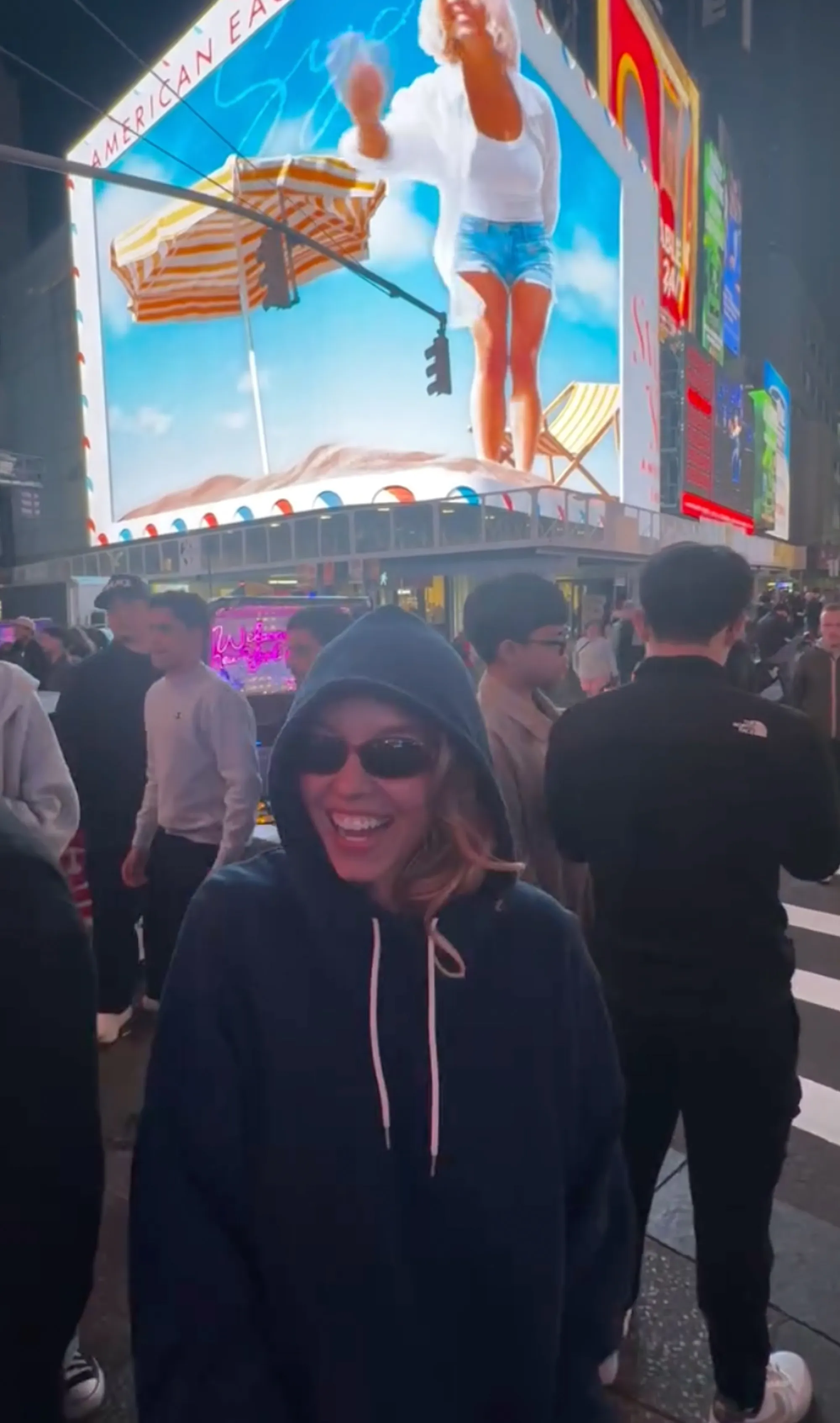 Sydney Sweeney in sunglasses and a hoodie, standing in Times Square with her American Eagle ad on a billboard behind her.