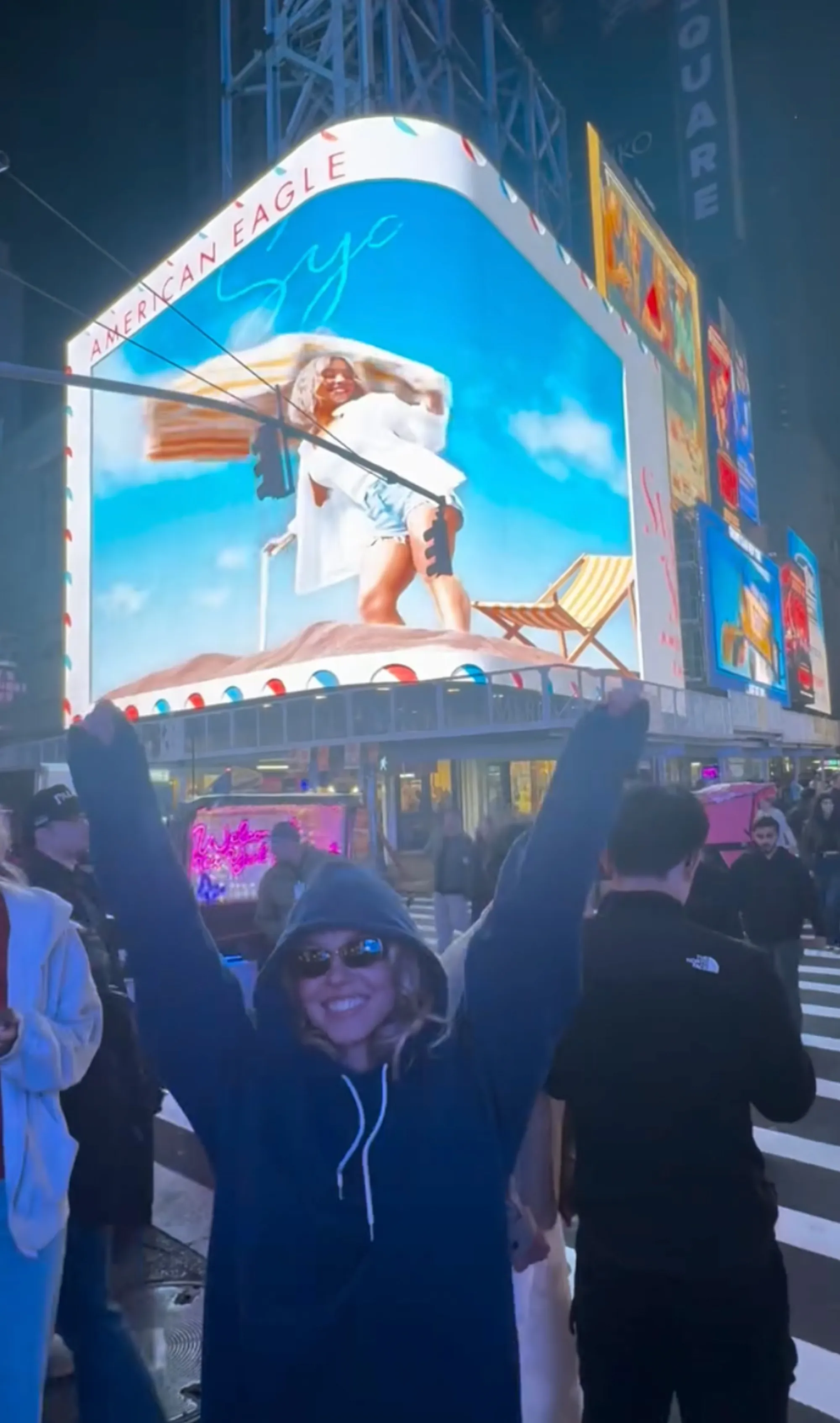 Sydney Sweeney raising her arms in celebration in front of a Times Square billboard featuring her American Eagle campaign.