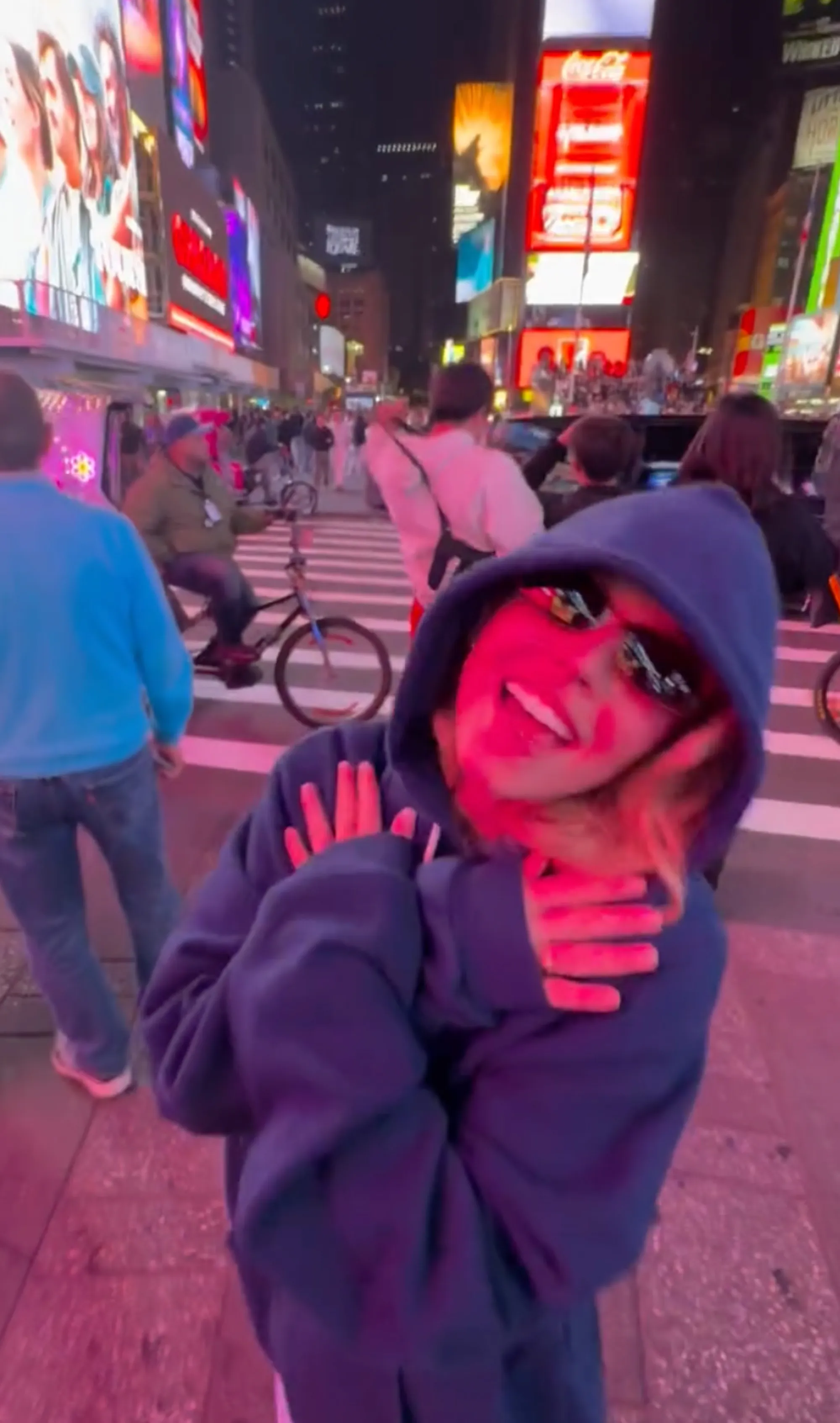 A person in a hoodie with pink light on their face, posing in Times Square.
