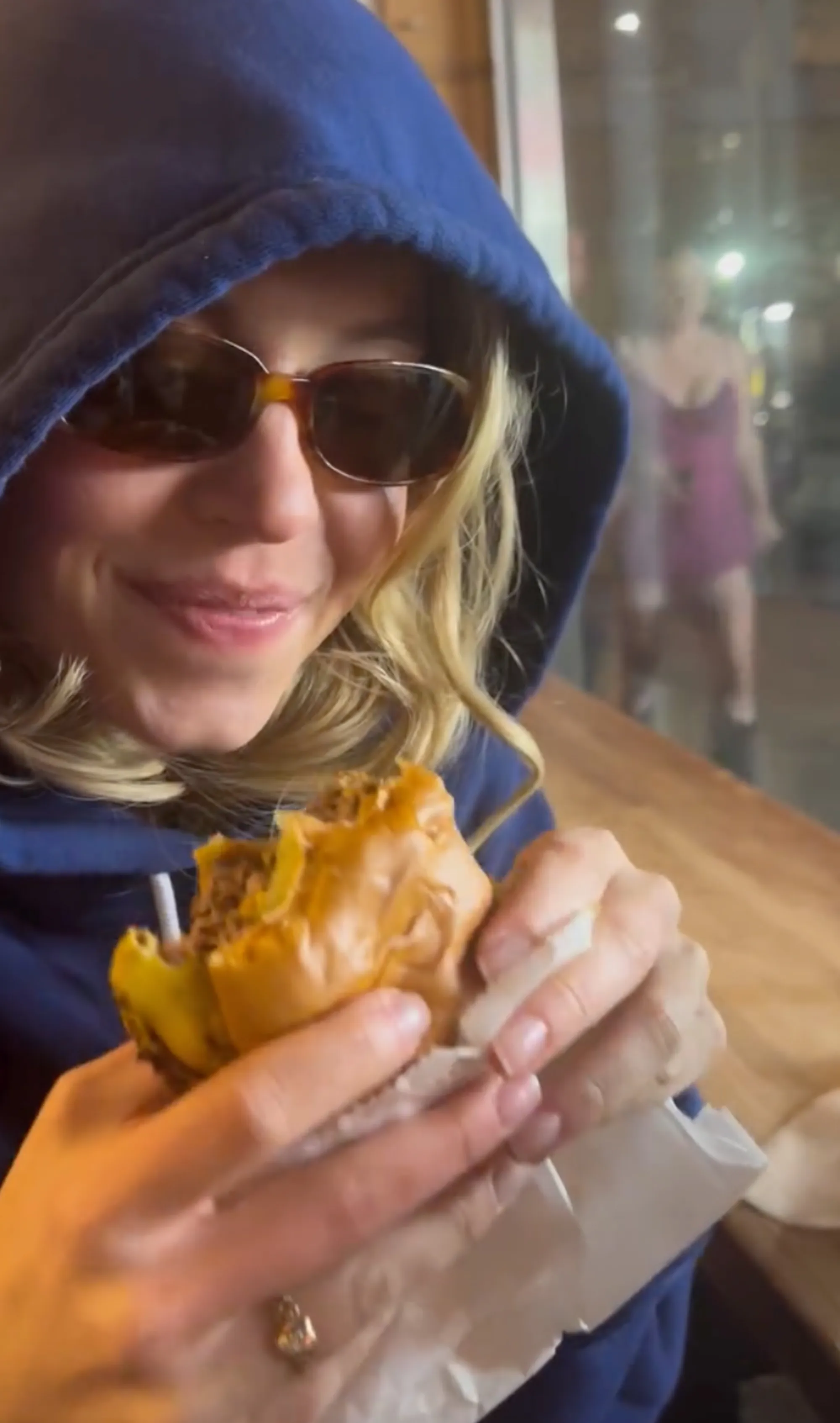 Sydney Sweeney eating a burger with sunglasses and a blue hoodie on.