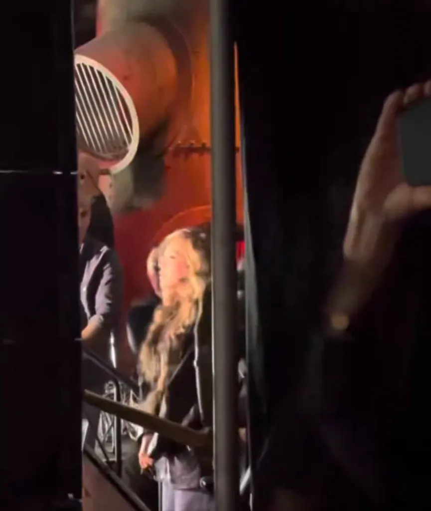 Beyoncé with long hair, wearing a black top and light pants, watching a performance in a dimly lit venue.