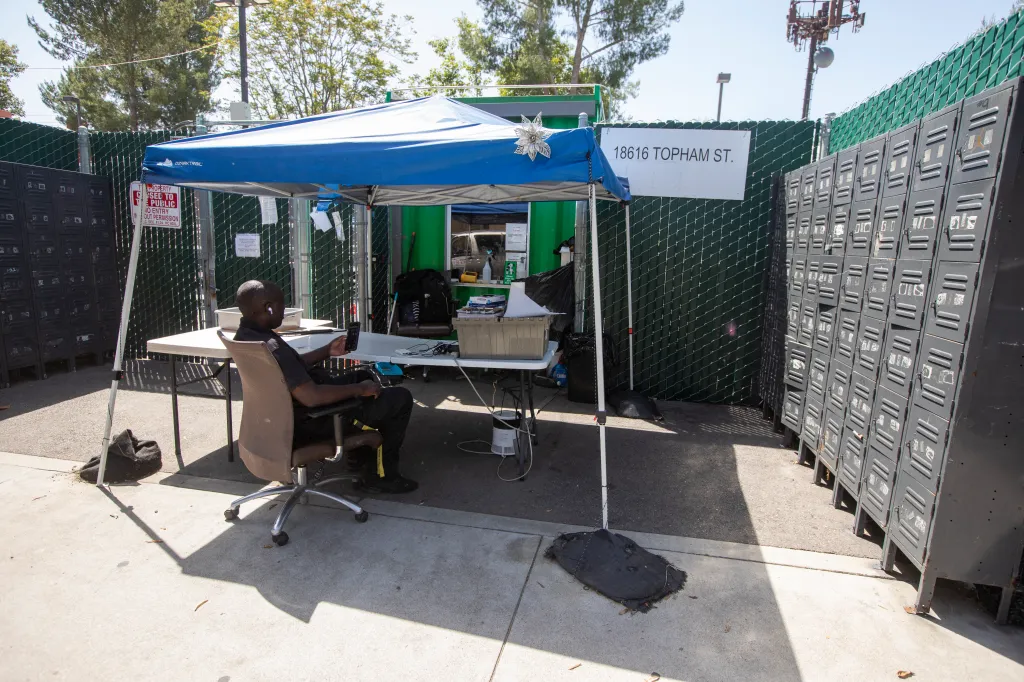 A security guard sits at a table under a canopy outside a homeless shelter.