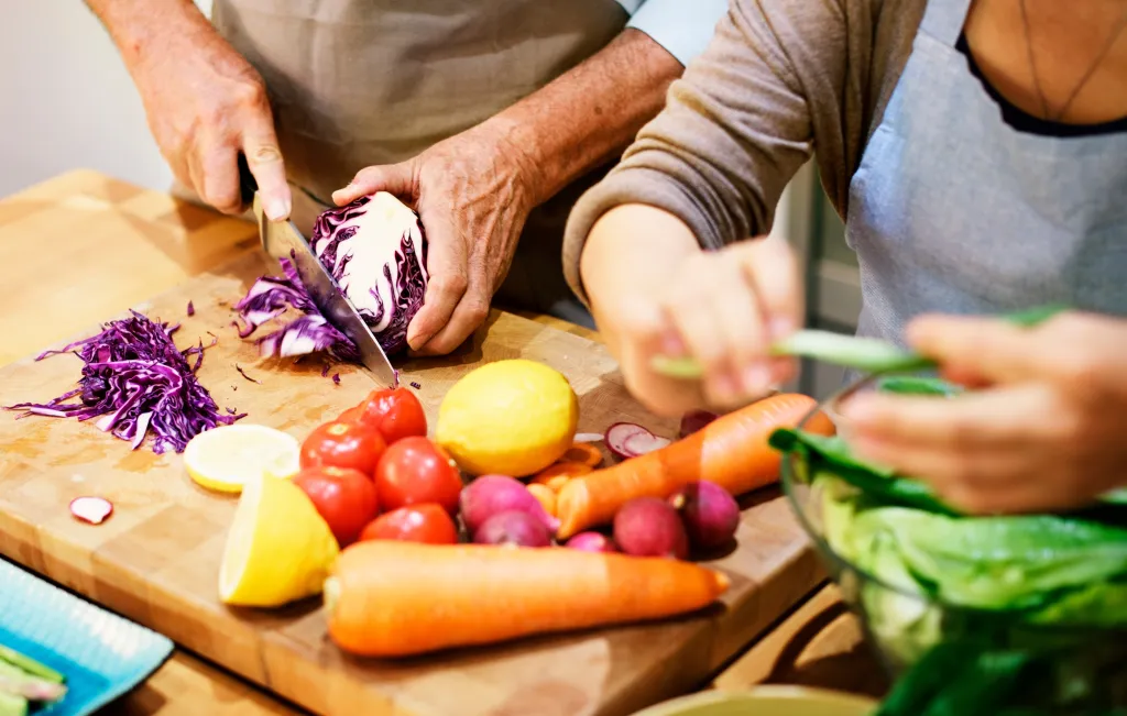 A man chopping red cabbage and a woman adding greens to a salad bowl.