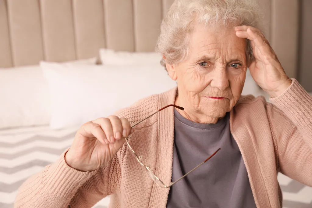 A senior woman with gray hair, dressed in a peach cardigan and gray shirt, sits on a bed with her hand on her head and glasses in the other hand, looking distressed.