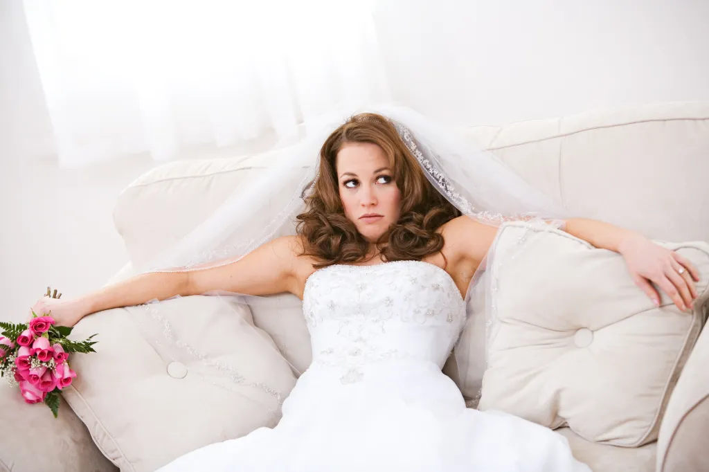 Upset bride in a white gown and veil sits on a couch, holding a pink rose bouquet.