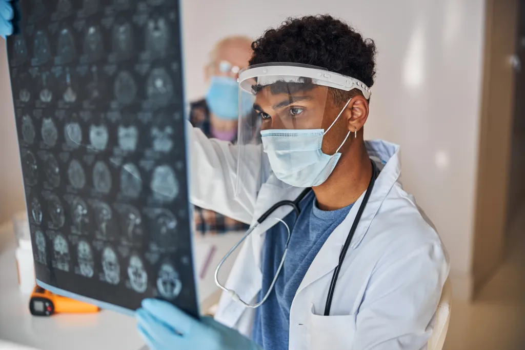 A neurologist wearing a face shield and mask examines brain MRI scans.