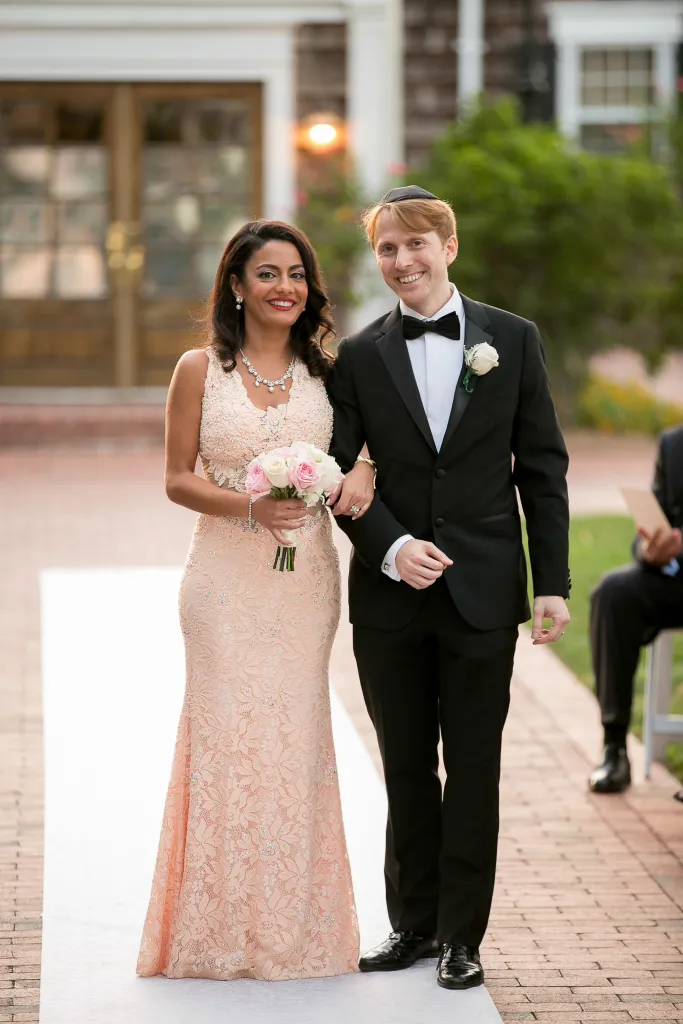 Seterah in a peach lace gown and Ryan in a black tuxedo walking down an aisle, both smiling.