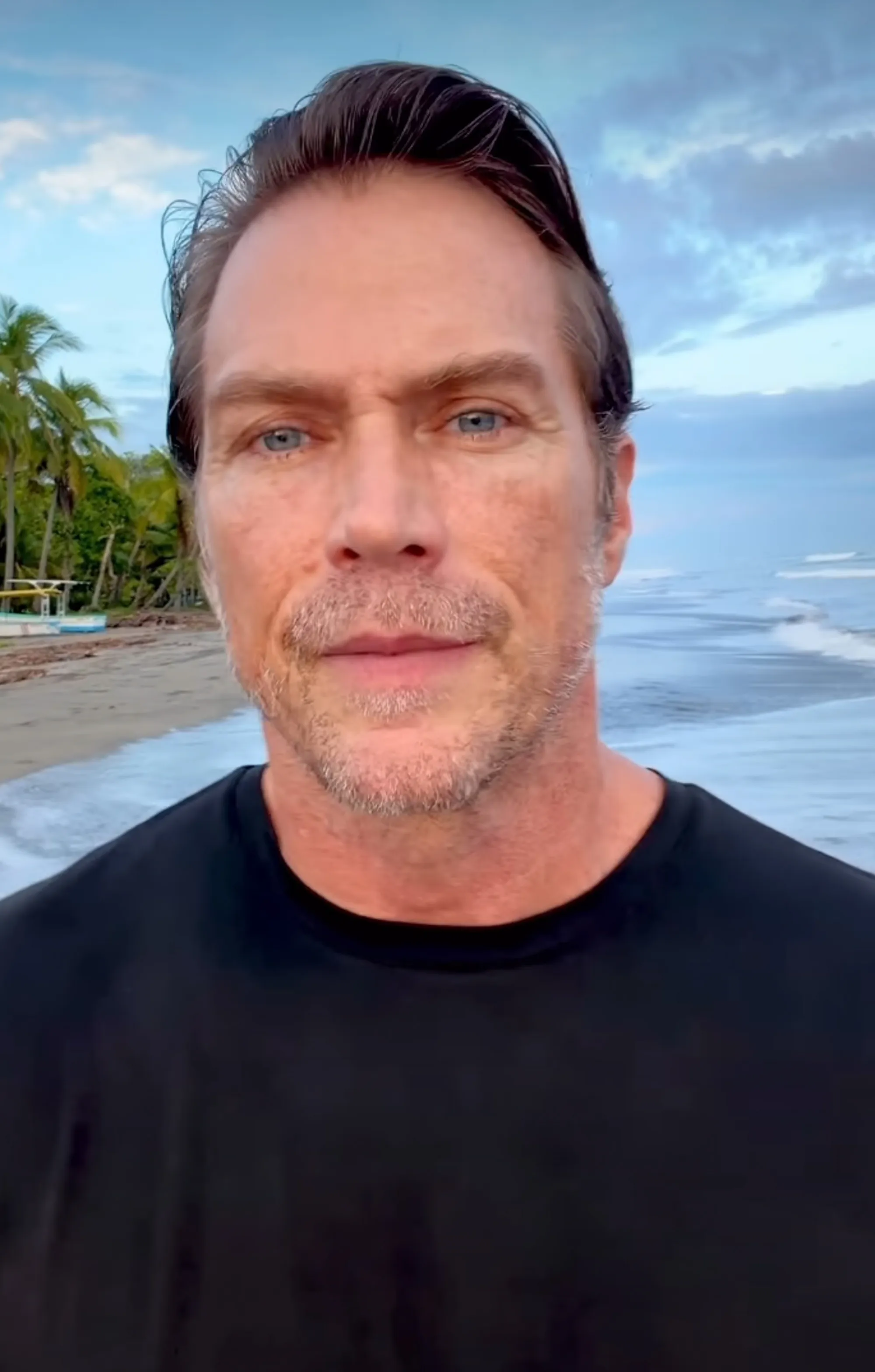 Jason Lewis on a beach with palms, ocean, and sky in the background.