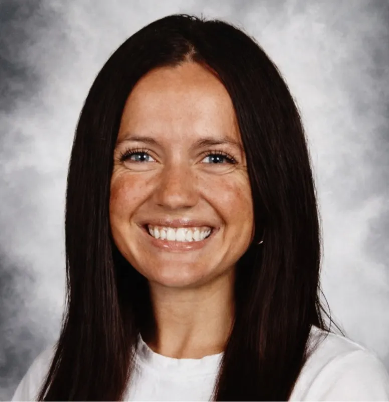 Headshot of a smiling woman with long dark hair, a white shirt, and a mottled grey background.