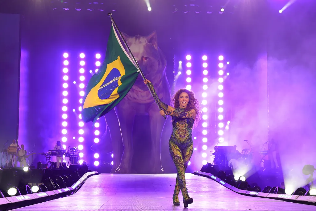Shakira on stage, holding a Brazilian flag, with a large wolf-like figure in the background and a band performing.