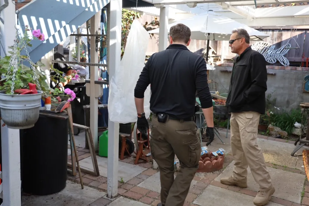 Two men standing outdoors in a paved area with various items around them, likely during an investigation.
