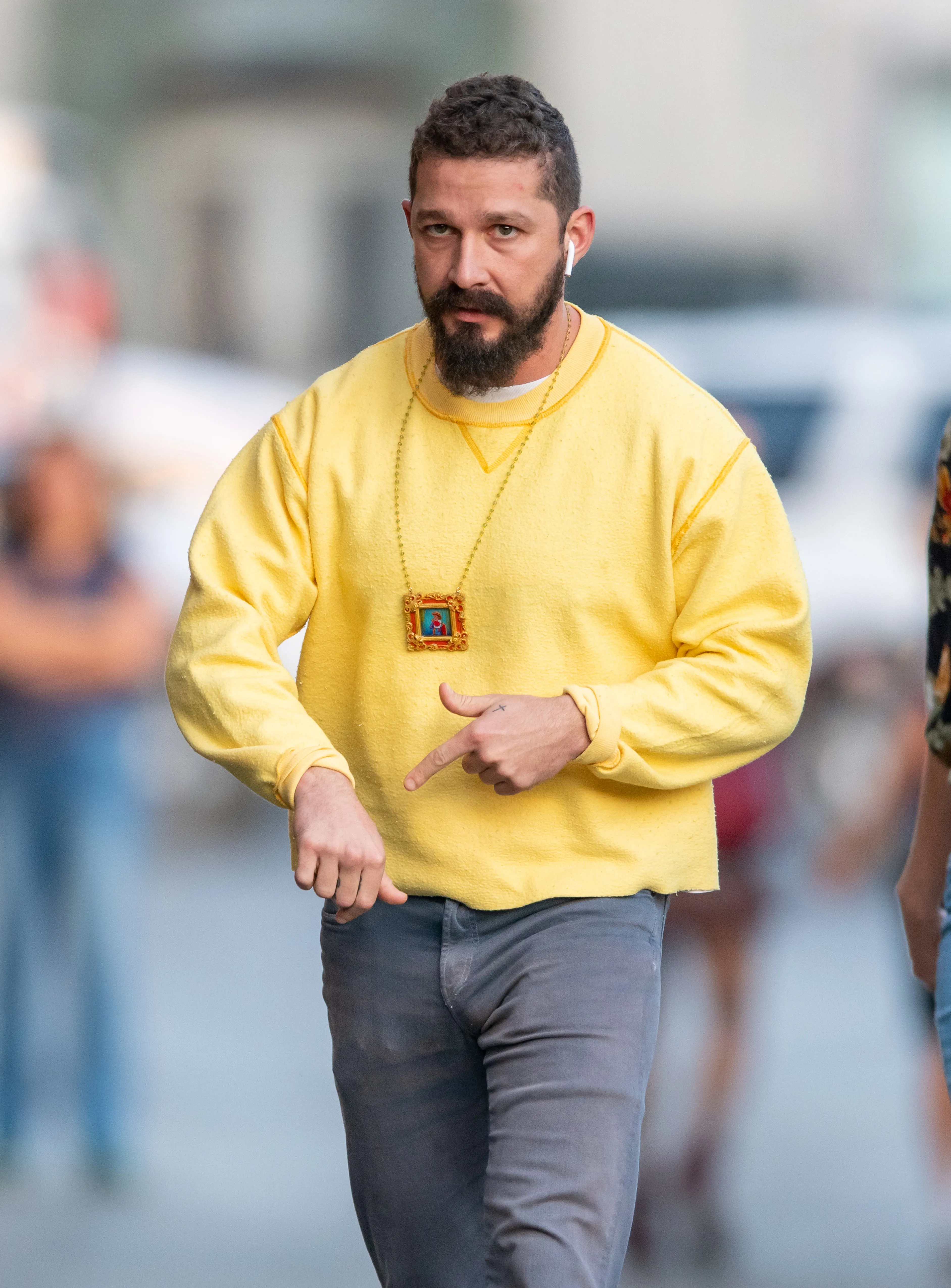 Shia LaBeouf wearing a yellow sweatshirt and gray pants, with a bearded face, AirPods, and a framed image on a necklace, points with his left hand.