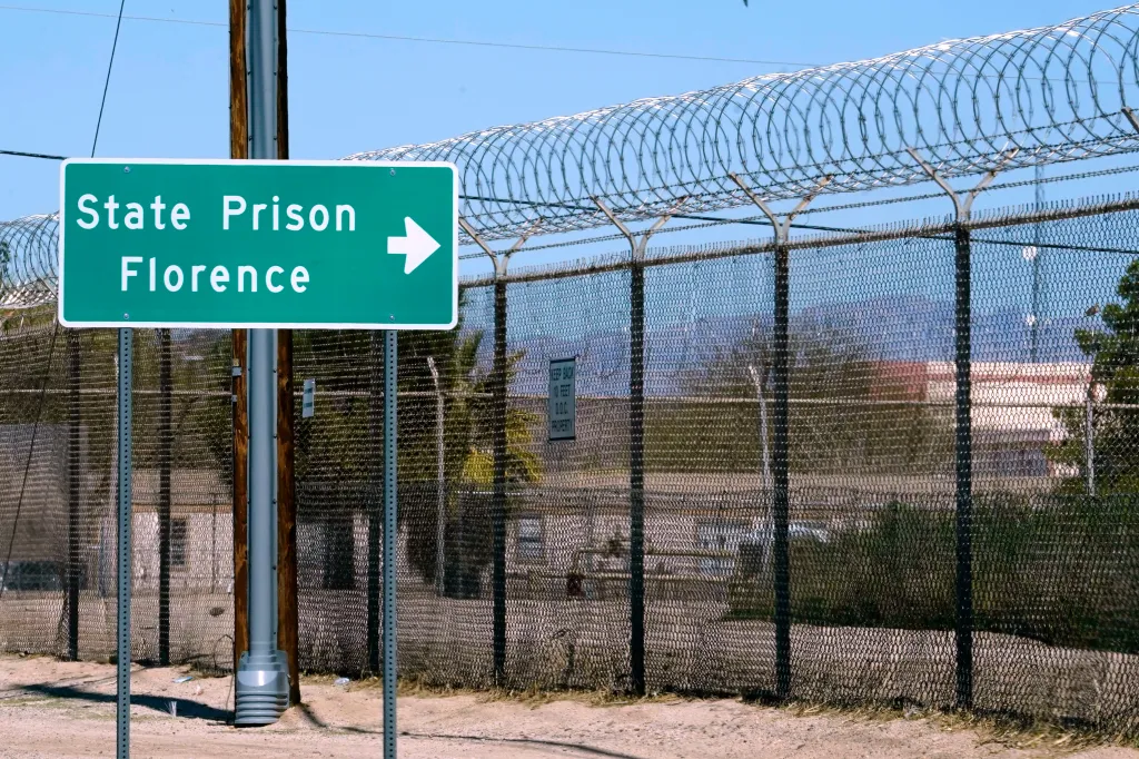 A sign for State Prison Florence with an arrow pointing right, next to a barbed wire fence.
