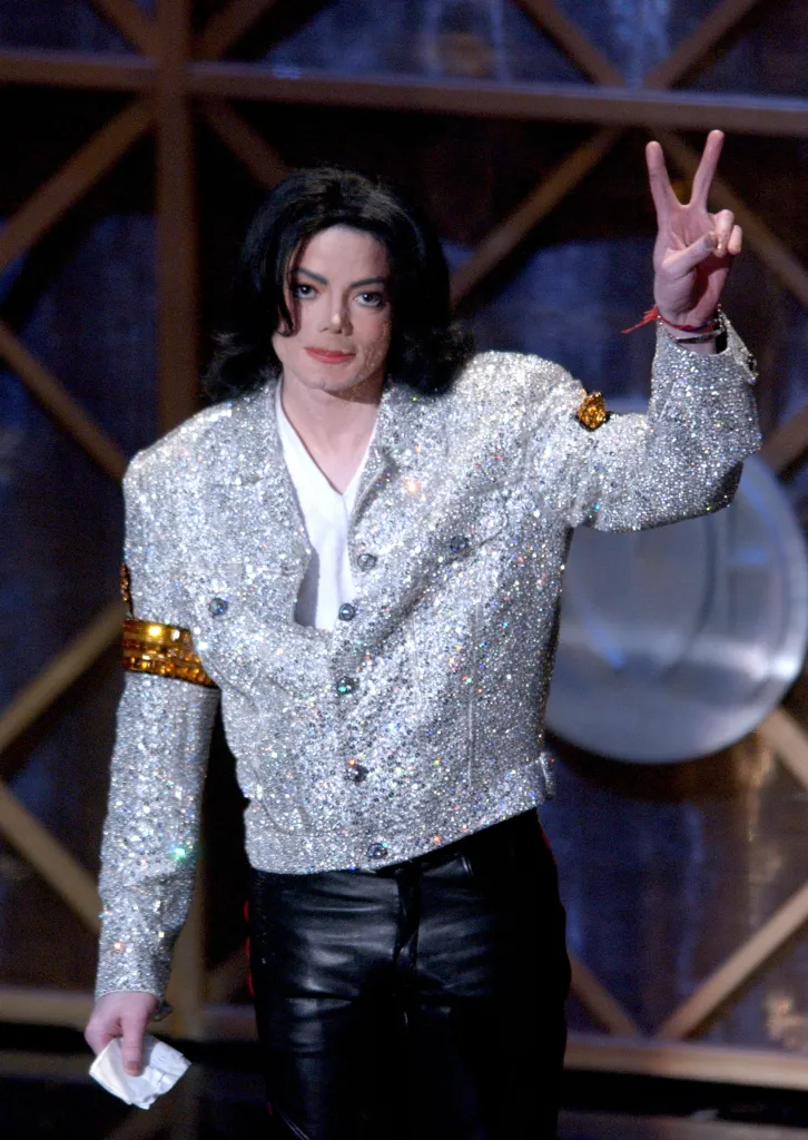 Michael Jackson holding a tissue and making a peace sign, wearing a sparkly silver jacket and black leather pants.