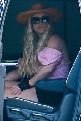 Britney Spears wearing a wide-brim hat, sunglasses, pink off-shoulder top, black shorts, and a ring, sitting in a car.