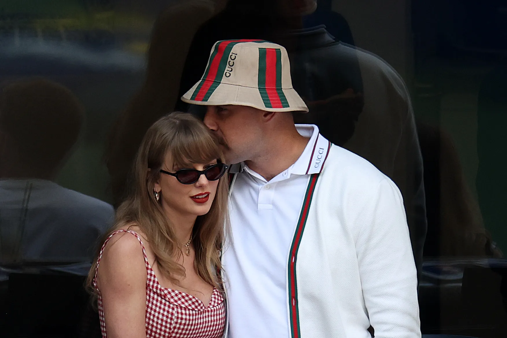 Taylor Swift in a red and white checkered top and Travis Kelce in a white polo, white cardigan, and Gucci bucket hat.