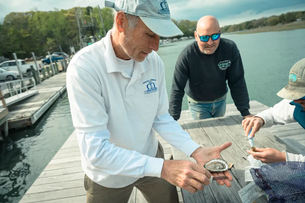 Sixto Portilla, Chief of Operations for Maris Stella, holding an oyster on a dock with another man shucking an oyster beside him.