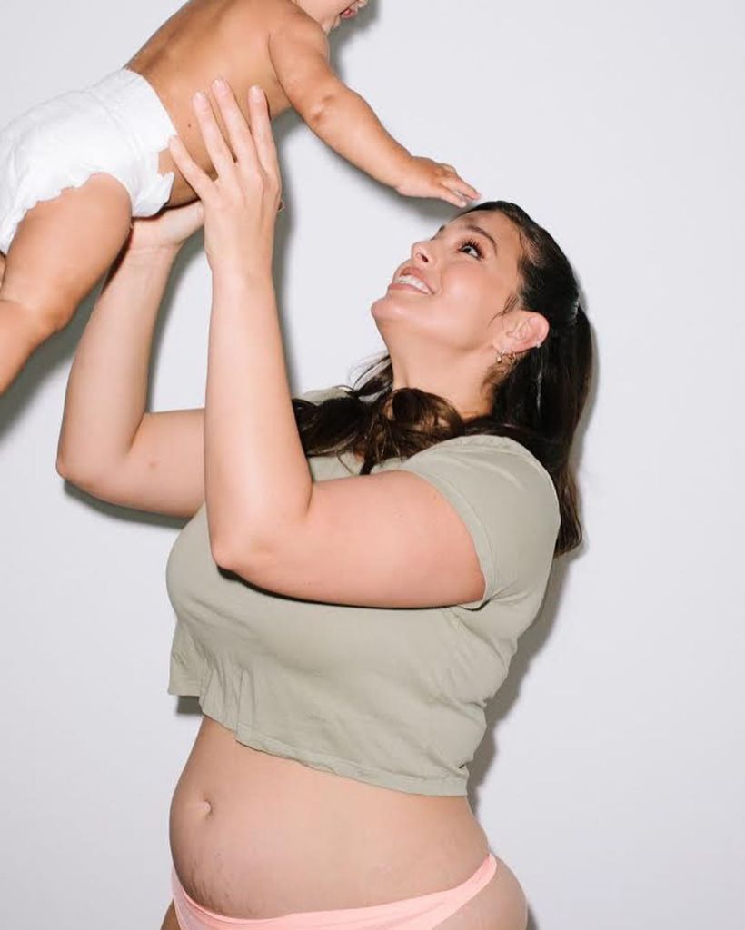 Ashley Graham holding her baby up, revealing her postpartum belly.