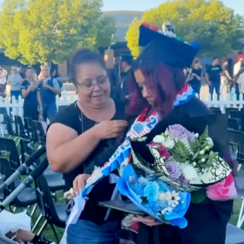 A woman in a graduation gown holds flowers while another woman adjusts her stole.