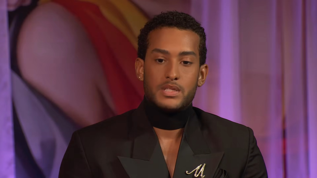 A man with dark curly hair, a black suit, and a letter M pin.