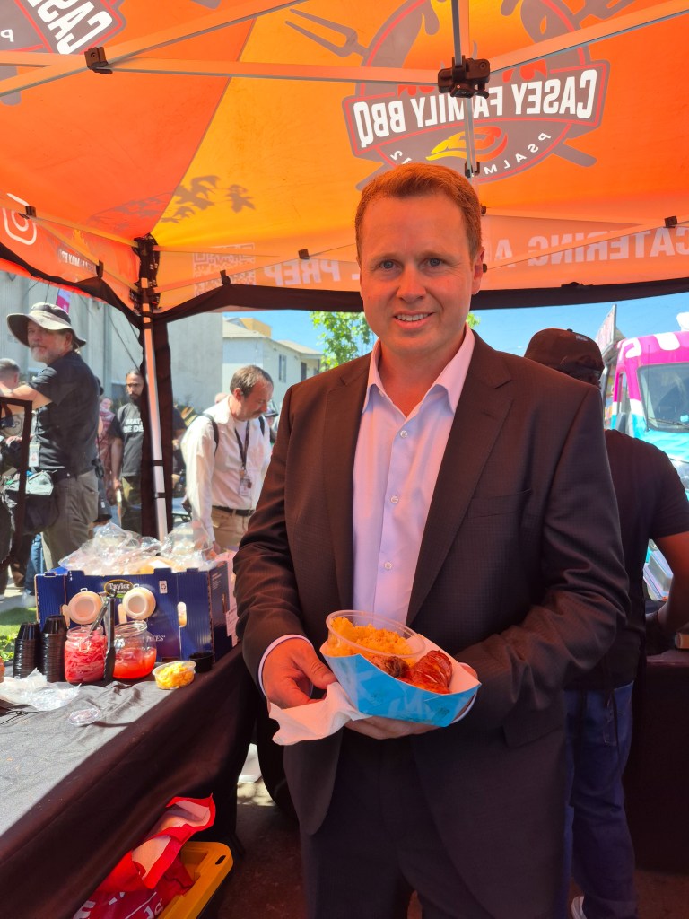 Spencer Pratt smiling and holding a container of BBQ food at a meet and greet event.