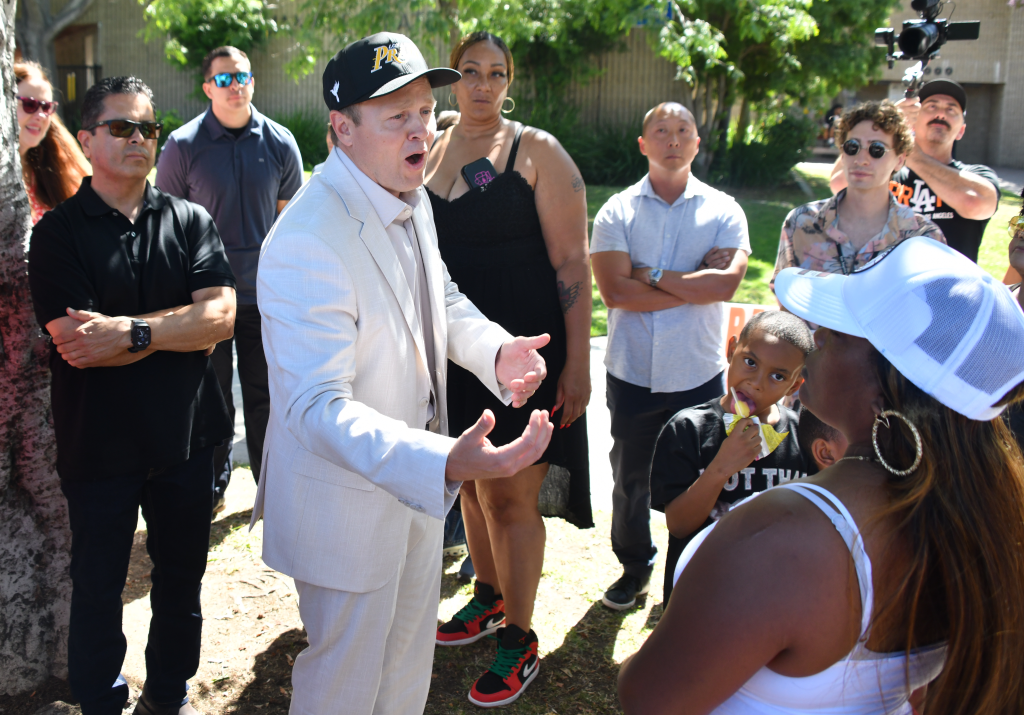 Spencer Pratt, wearing a black cap, white suit, and light gray tie, gestures with open hands while speaking to a group of people, including African American and Latino voters, in Tim Gilliam Park in Los Angeles.