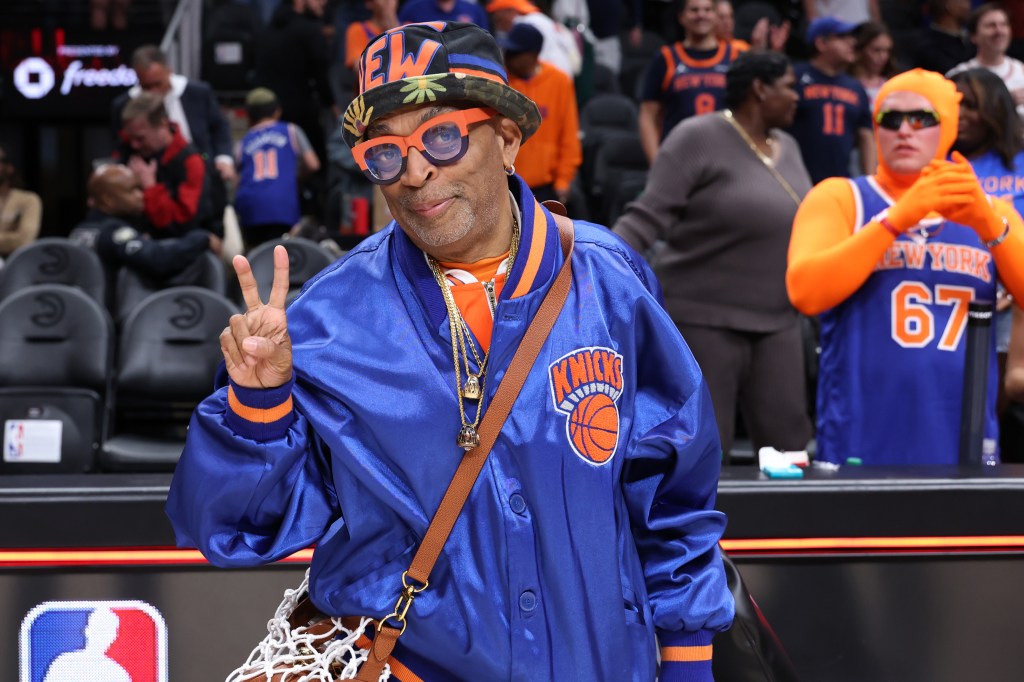 Spike Lee in a New York Knicks jacket and hat, making a peace sign at a basketball game.