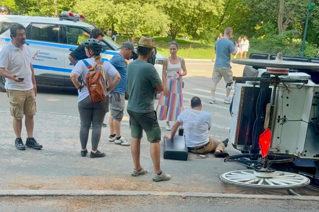 A group of people standing around an overturned horse carriage, with one person sitting on the ground, after a horse-drawn carriage accident.