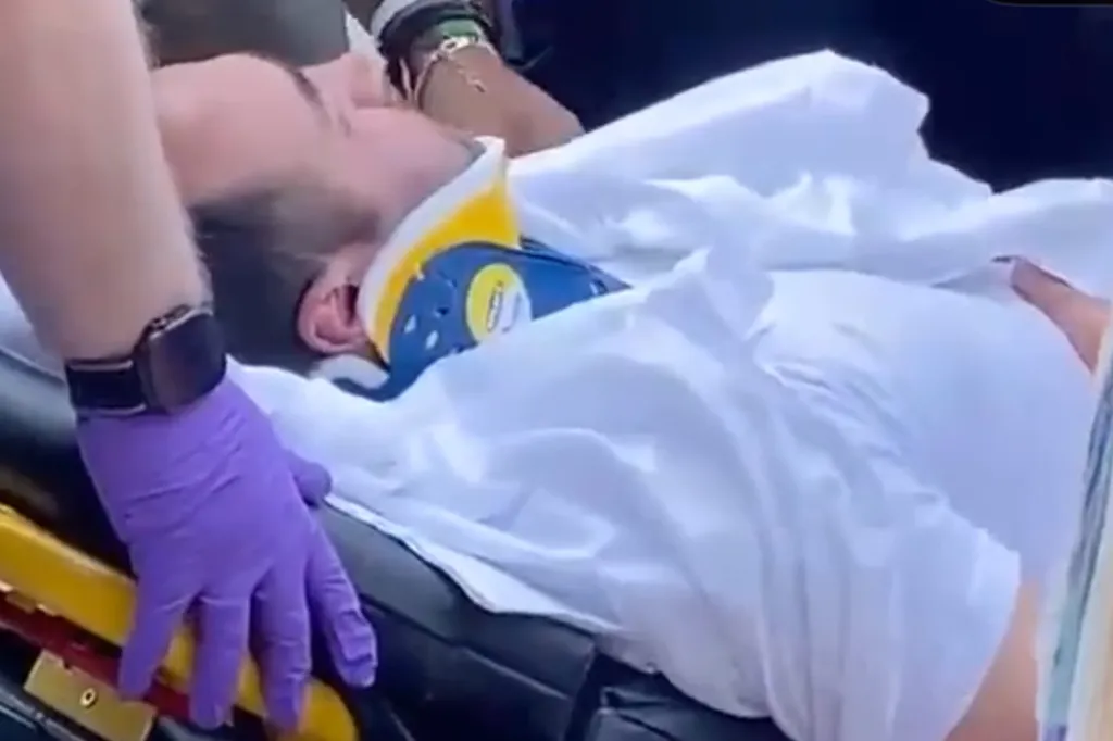 A person wearing a neck brace lies on a stretcher, being attended to by someone in purple gloves.