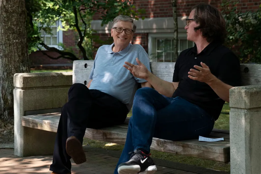 Bill Gates smiling on a bench next to a man gesturing during an interview.