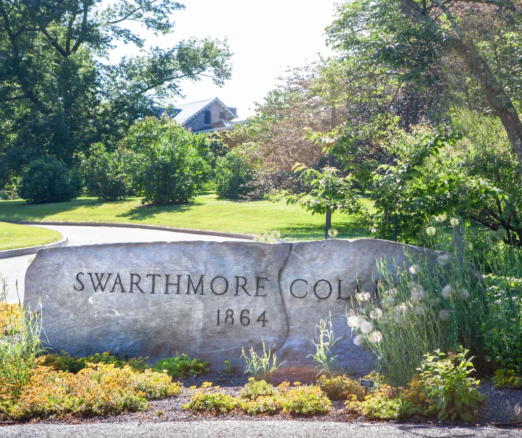 Stone sign for Swarthmore College, founded in 1864, surrounded by lush greenery and a paved road leading into the campus.