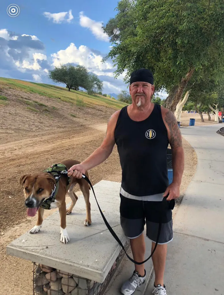 Darrell Sheets smiling with his dog on a leash, standing on a walking path with a grassy hill and trees behind them.