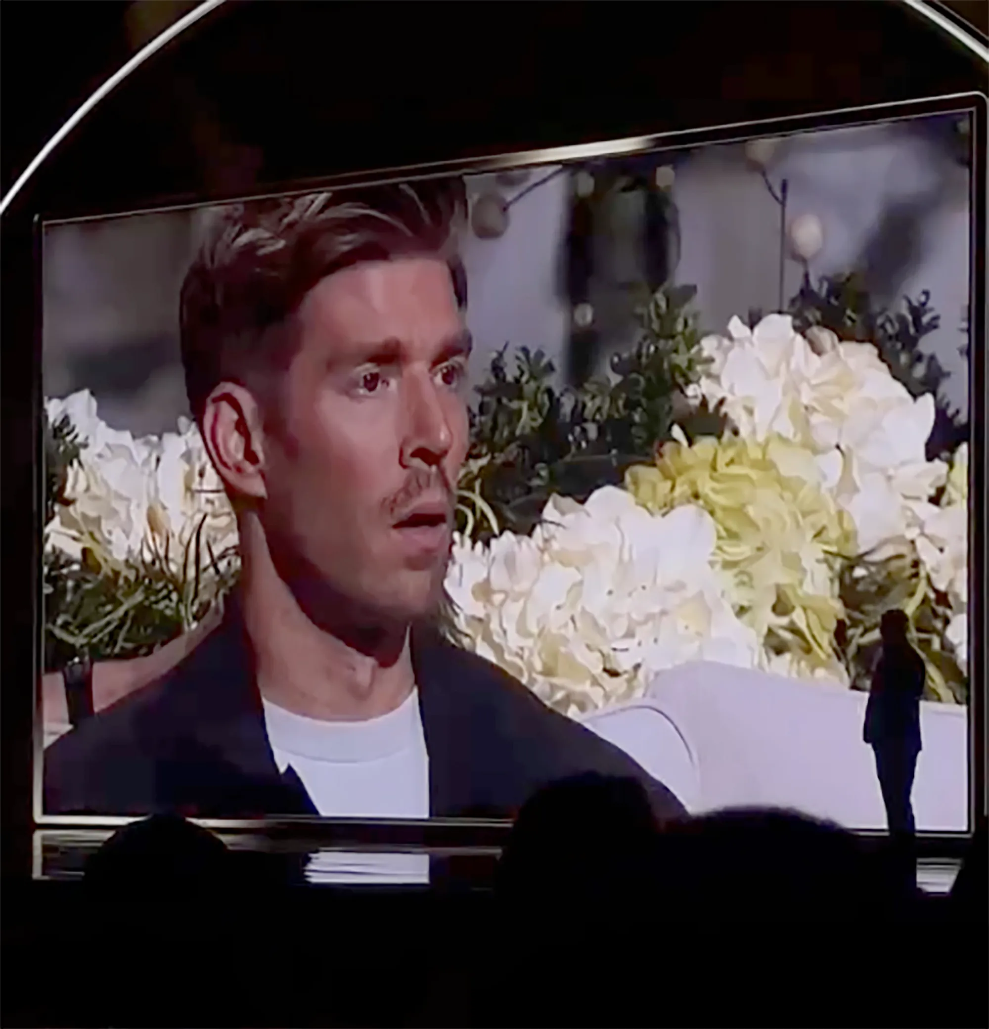 Man with short hair and a mustache on a screen, with a white couch and flowers behind him.