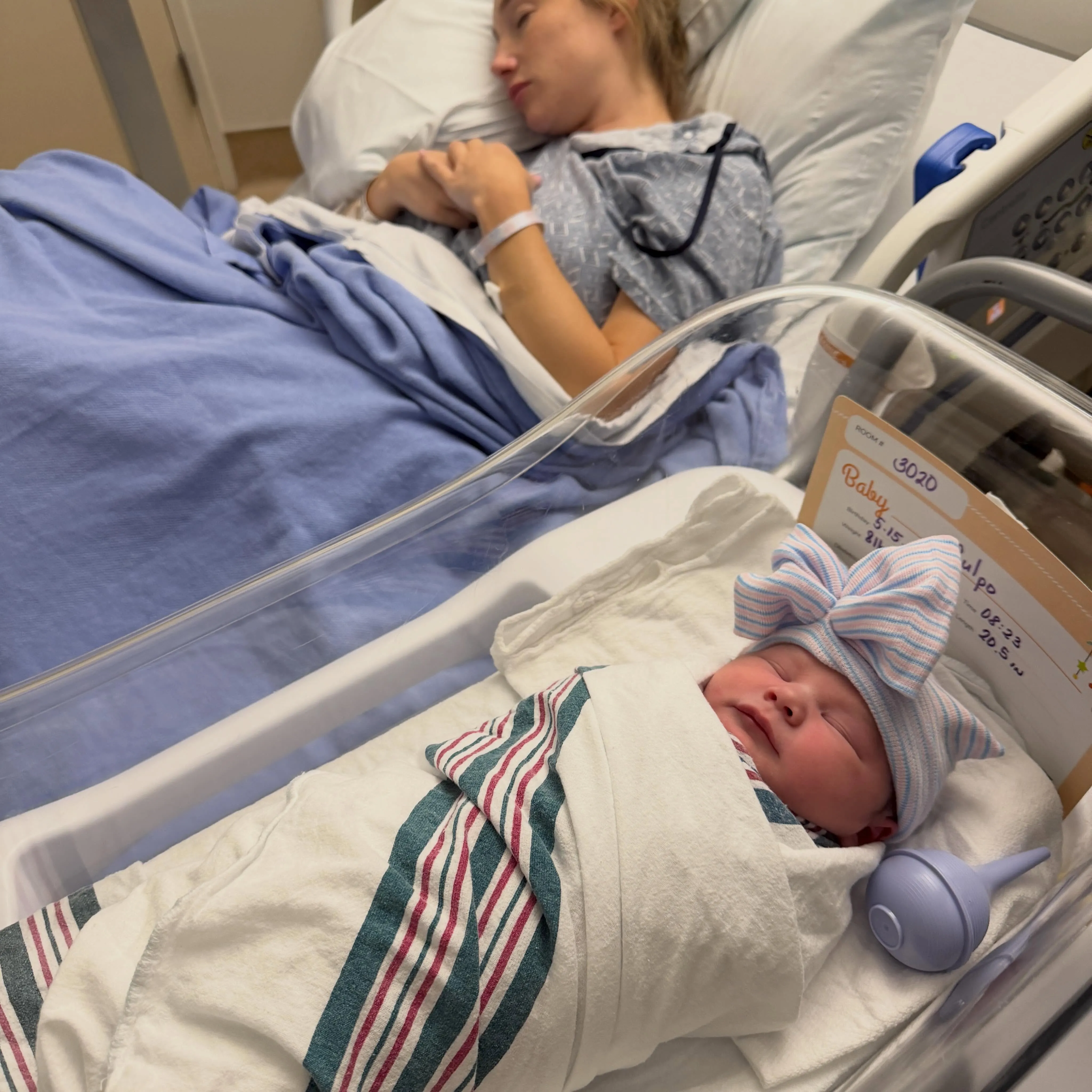 A newborn baby sleeps in a bassinet next to its mother who is also sleeping in a hospital bed.