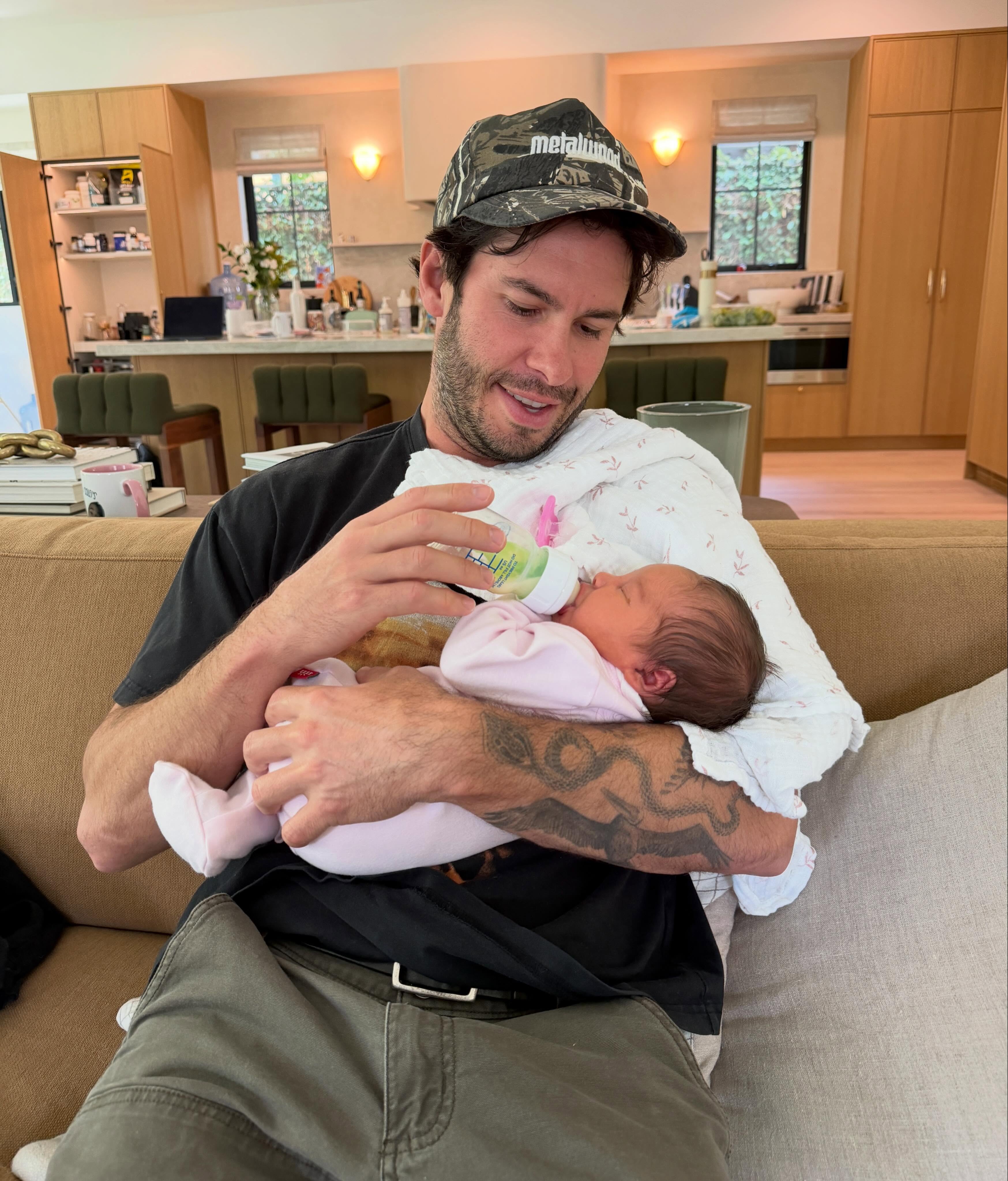A man with a camouflage hat and tattoos on his arm feeds a baby from a bottle while sitting on a sofa.