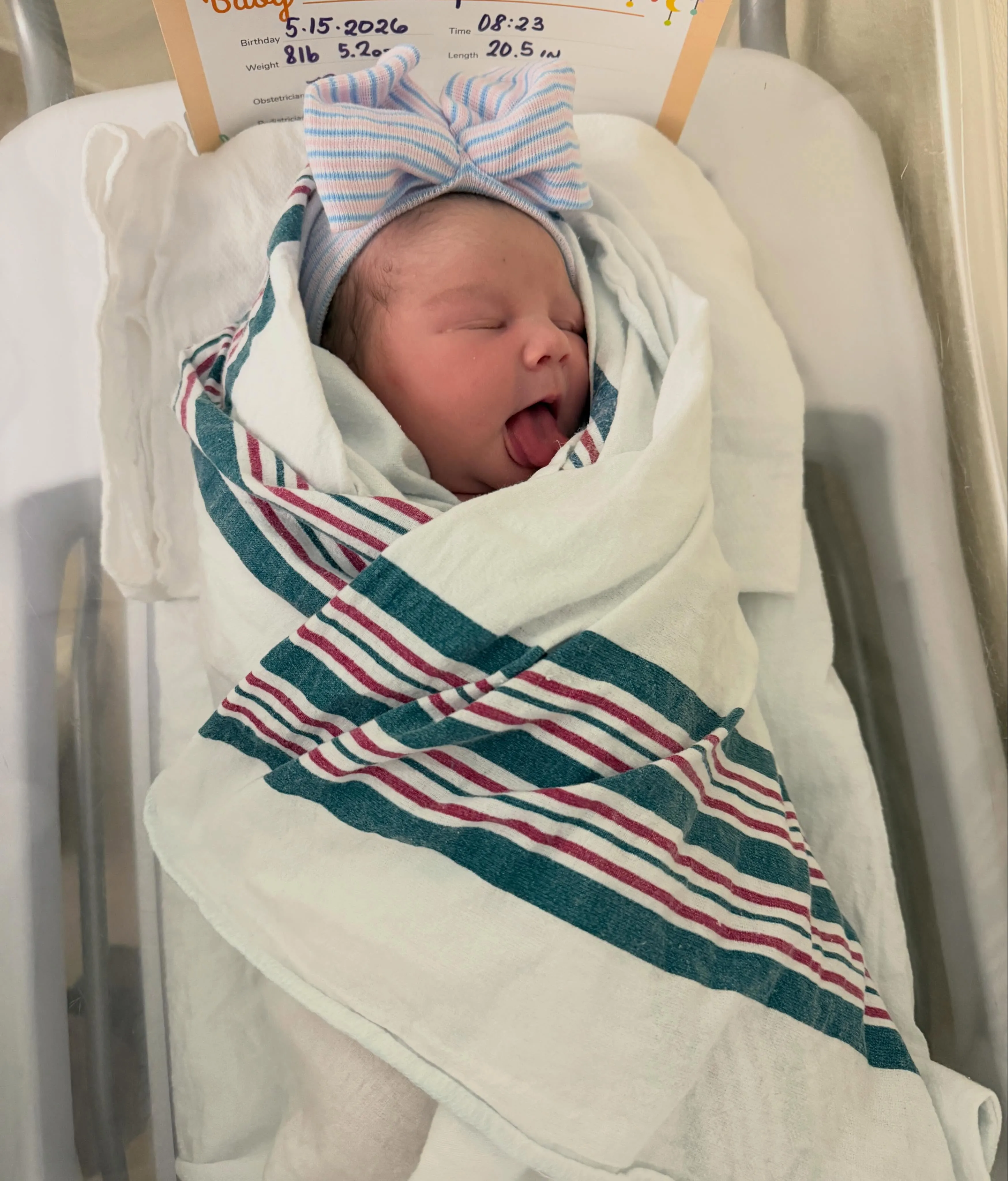 A newborn baby, Selene, wrapped in a white blanket with blue and red stripes, wearing a pink and blue striped bow-shaped hat, with her tongue sticking out.