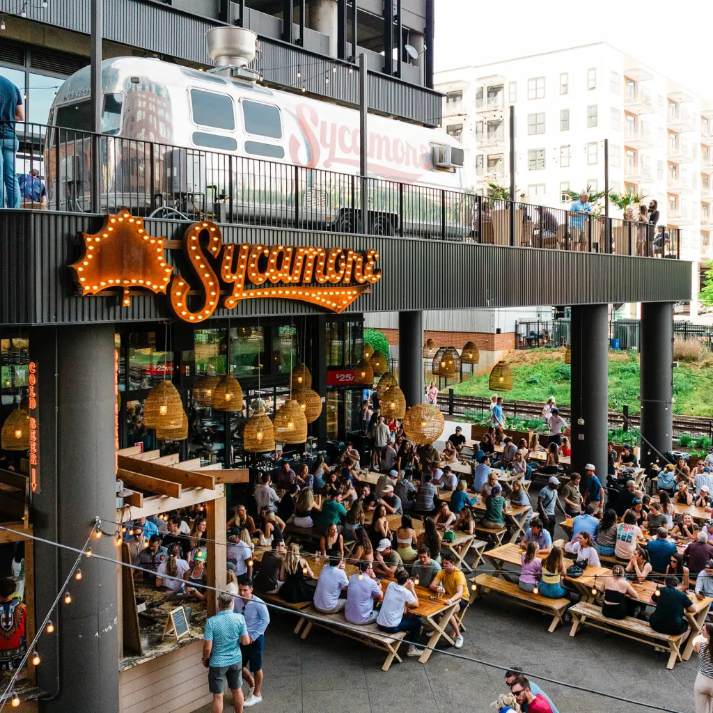 The exterior of the Sycamore Taproom, with a large illuminated sign and an Airstream trailer on a balcony above a crowded outdoor seating area.