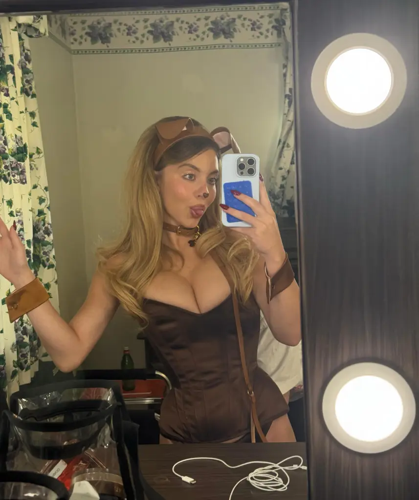 Sydney Sweeney wearing a brown corset, dog ears, and a choker while taking a mirror selfie and sticking out her tongue.