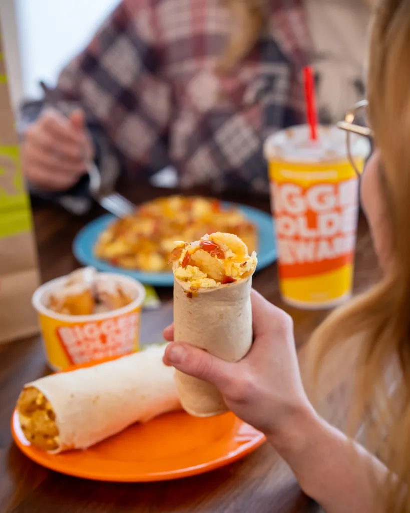 A person holding a Taco John's breakfast burrito, with another burrito, a side of hash browns, and a drink on the table.
