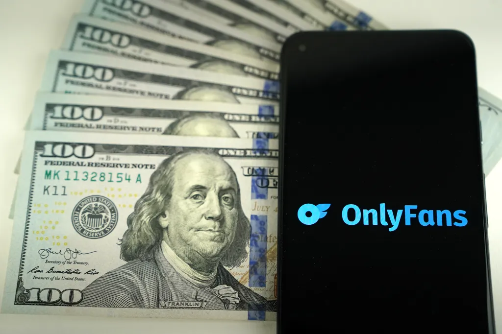 OnlyFans logo on a smartphone screen next to a pile of US one-hundred dollar bills.