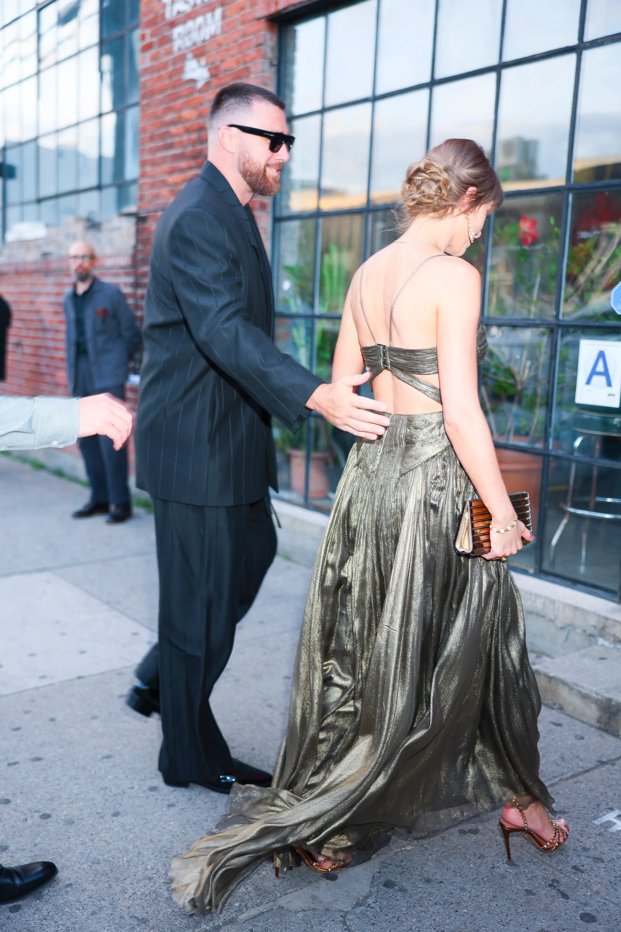 Taylor Swift in a gold dress with Travis Kelce in a pinstripe suit, walking together.