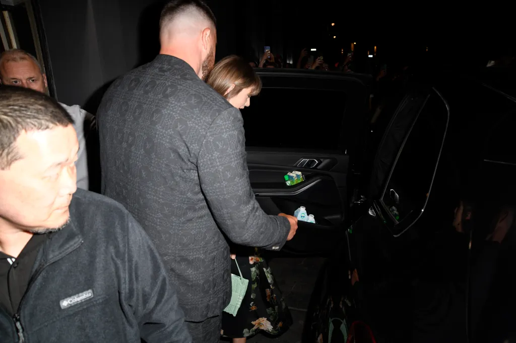 Taylor Swift, in a black floral dress and green handbag, and Travis Kelce, in a grey suit, entering a car with spectators visible in the background.