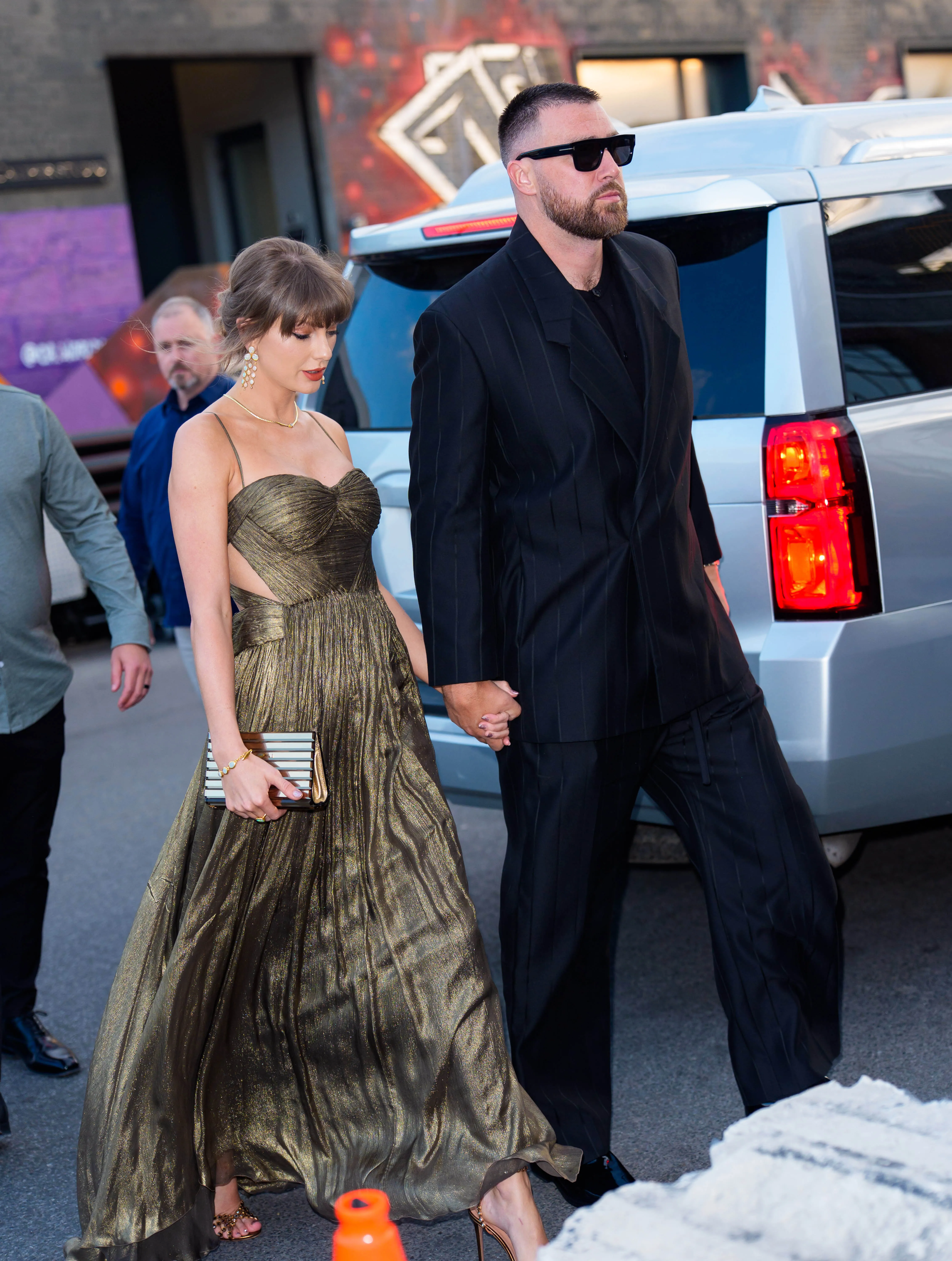 Taylor Swift and Travis Kelce holding hands while walking, with Swift in a gold dress and Kelce in a pinstripe suit and sunglasses.