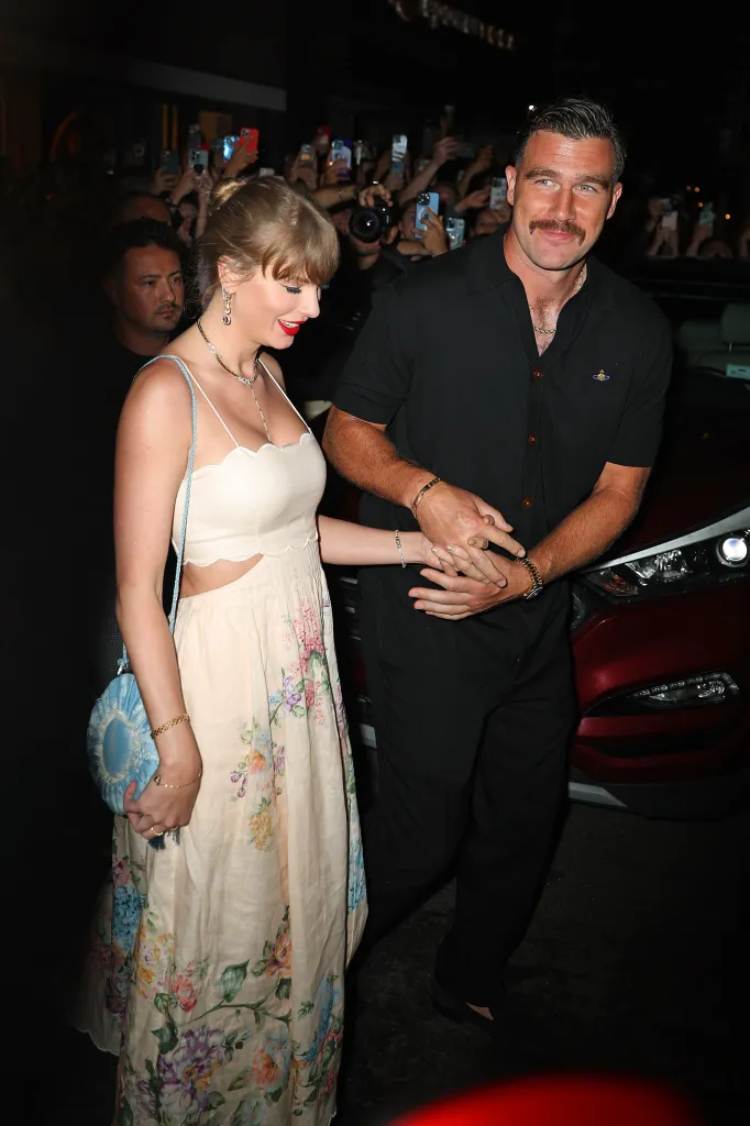 Taylor Swift and Travis Kelce holding hands while walking through a crowd of people taking photos.