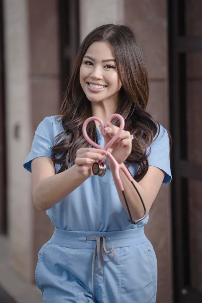 A smiling nurse in light blue scrubs holds up a pink stethoscope shaped into a heart.