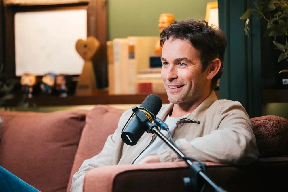 Chace Crawford sits on a couch in front of a microphone, smiling.