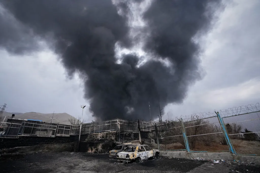 A thick plume of smoke rises from an oil storage facility in Tehran, Iran, with two burned-out cars in the foreground.